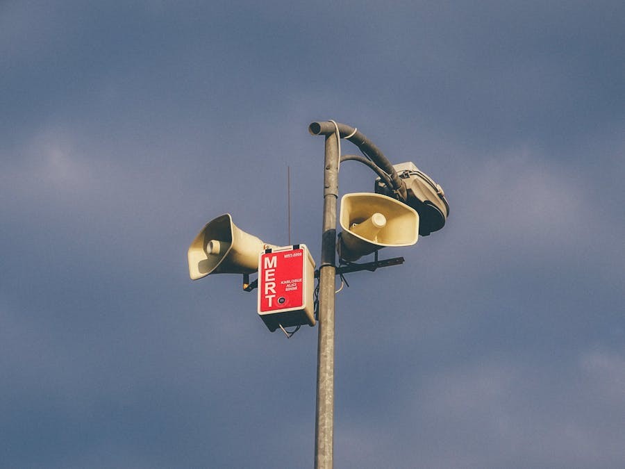 The image shows a tall pole with multiple loudspeakers mounted on it. A red mass notification unit with the label "MERT" is attached to the pole, positioned near the speakers. The sky in the background appears slightly cloudy.