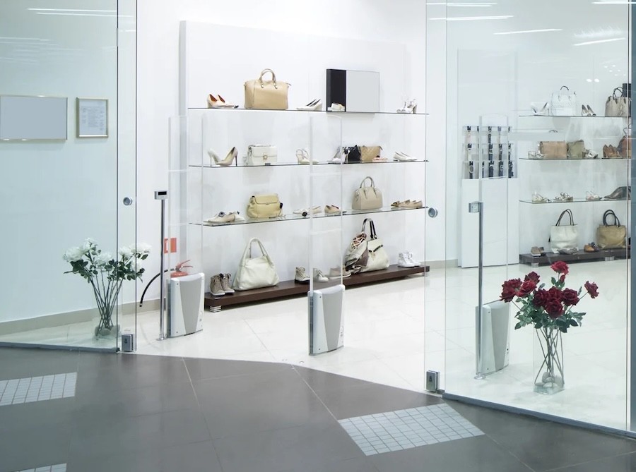 Luxury retail store with glass walls, handbags and shoes on display shelves, and security sensors at the entrance.