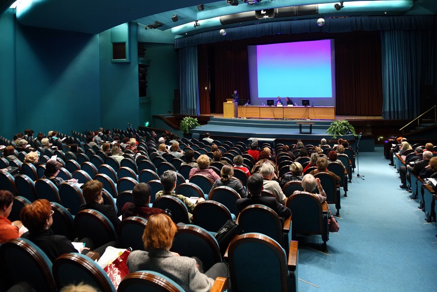 An audience seated in an auditorium watching a presentation on stage with a large projection screen.